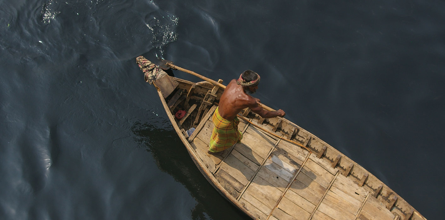 <p>A man in traditional <i>longi</i> paddles his sampan boat across a river.</p>